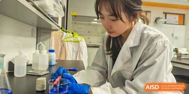 A focused female student wearing a white lab coat and blue protective gloves carefully conducts a chemistry experiment with a beaker in a modern science lab at Beijing Chaoyang KaiWen Academy.