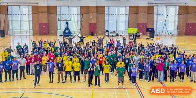 A large group of students and faculty members in colorful team shirts gather for a massive group photo on the indoor basketball court at Beijing Chaoyang KaiWen Academy.