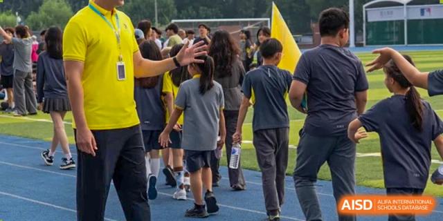 Students in grey and yellow uniforms walk along the blue athletic track during an outdoor sports day event under the supervision of a teacher at Beijing Chaoyang KaiWen Academy.