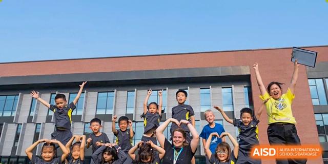 A group of young elementary students poses with their teacher in front of a modern school building while making heart shapes with their hands at Beijing Chaoyang KaiWen Academy.