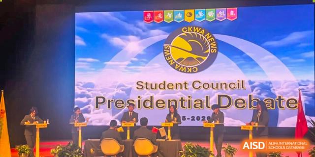 Student council candidates sit at a formal table for a presidential debate in front of a large digital screen featuring the CKWA News logo at Beijing Chaoyang KaiWen Academy.