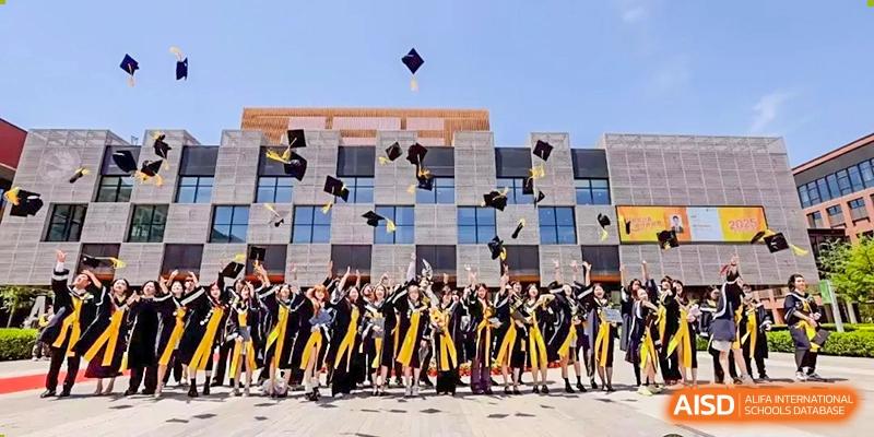 A large group of graduates in traditional black and yellow academic regalia joyfully toss their caps into the air in front of a modern building at Beijing Chaoyang KaiWen Academy.