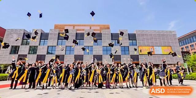 A large group of graduates in traditional black and yellow academic regalia joyfully toss their caps into the air in front of a modern building at Beijing Chaoyang KaiWen Academy.