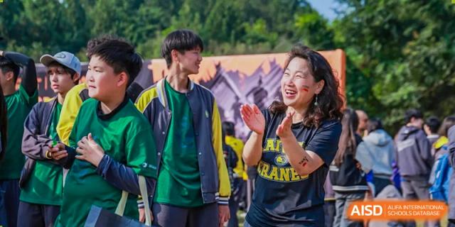 A female staff member in a black t-shirt claps enthusiastically while cheering for students during an energetic outdoor campus event at Beijing Chaoyang KaiWen Academy.