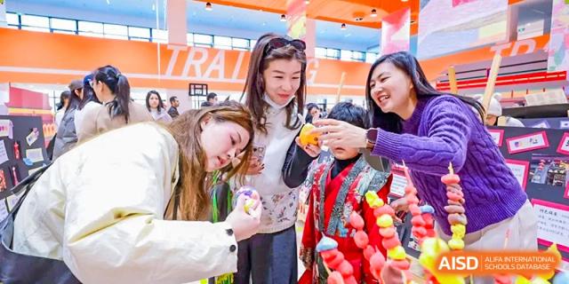 Two teachers smile while helping a young student in a traditional red outfit explore colorful cultural crafts during a school festival at Beijing Chaoyang KaiWen Academy.
