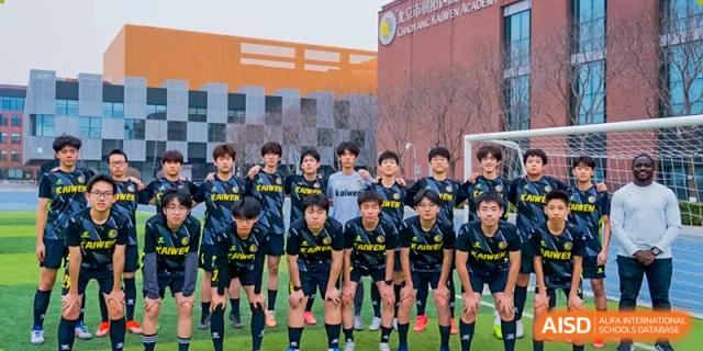 A youth soccer team in matching black and yellow jerseys poses together for a formal group photo on a green field in front of a modern campus building at Beijing Chaoyang KaiWen Academy.