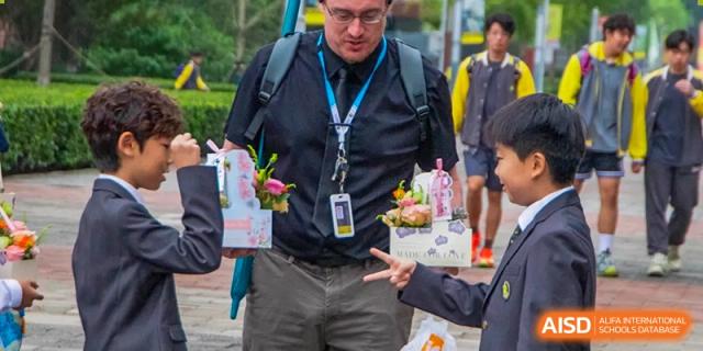 A teacher in a black shirt and glasses receives flower boxes from students during a special appreciation event held outdoors on the school campus at Beijing Chaoyang KaiWen Academy.