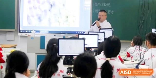 A teacher uses a microphone to lead an interactive lesson in front of a digital screen while students in white uniforms listen attentively at RDFZ Sanya School