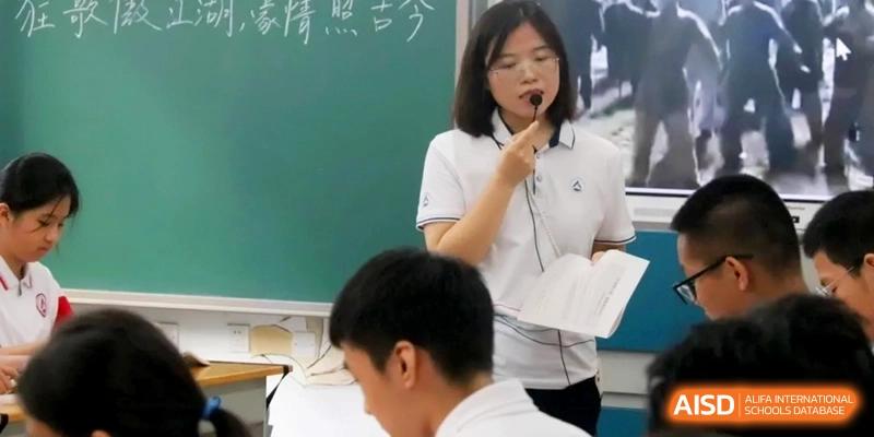 An educator holds a notebook and speaks into a microphone while leading a classroom activity for a group of seated students at RDFZ Sanya School