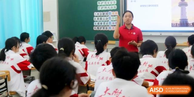 A teacher in a red polo shirt stands at the front of a classroom during a lecture while students take notes at their desks at RDFZ Sanya School