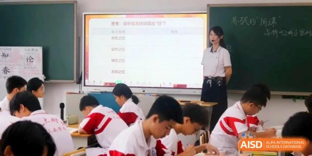 A teacher stands beside a digital screen displaying educational content while students in uniforms work at their desks in a modern classroom at RDFZ Sanya School