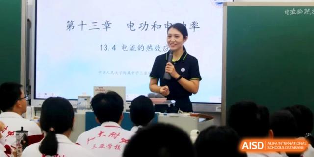 A smiling instructor holds a microphone and presents a lesson on electricity using a digital display in a bright classroom setting at RDFZ Sanya School