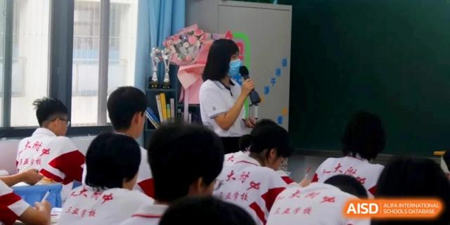 A teacher wearing a face mask speaks into a microphone while leading a classroom activity surrounded by trophies and educational materials at RDFZ Sanya School