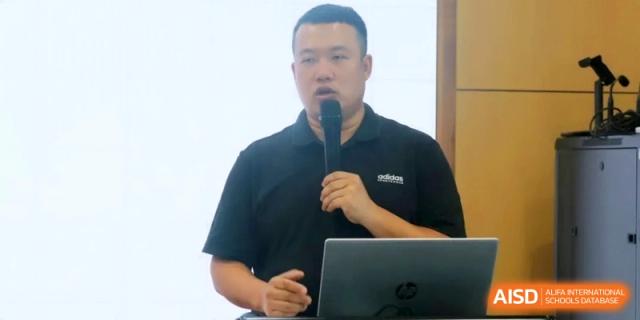 A man in a black polo shirt speaks into a microphone while standing behind a laptop during a formal presentation or lecture at RDFZ Sanya School