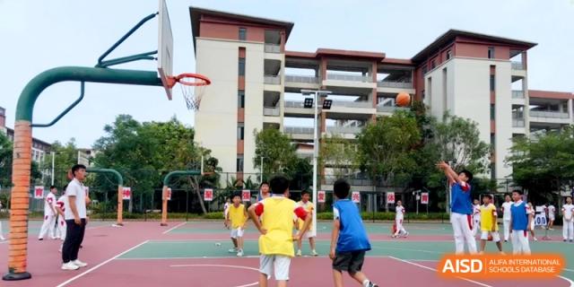 Young students in yellow and blue jerseys play a basketball game on an outdoor court with modern school buildings in the background at RDFZ Sanya School