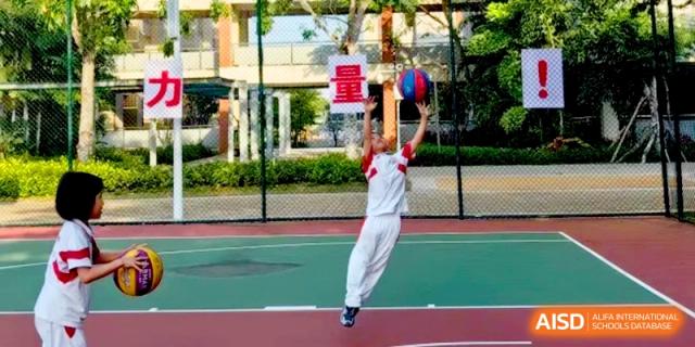 A student in a white and red uniform jumps high to shoot a basketball on an outdoor court surrounded by lush greenery at RDFZ Sanya School