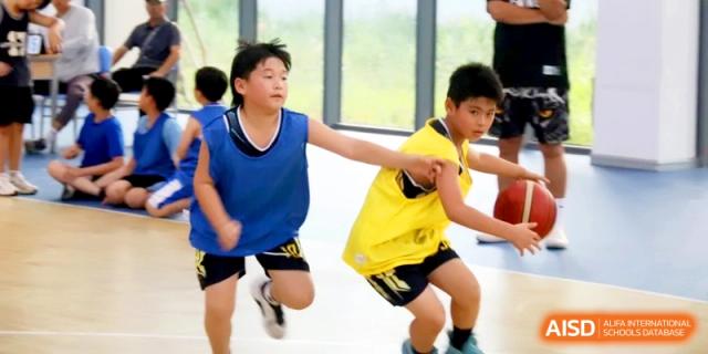 Two young boys in basketball uniforms compete for the ball during an energetic indoor sports match at RDFZ Sanya School