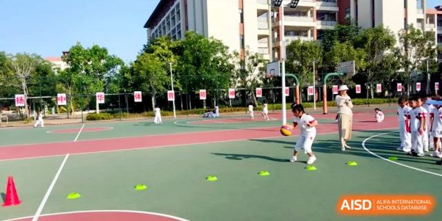 A student in a white uniform practices basketball drills by dribbling around green cones on an outdoor sports court at RDFZ Sanya School