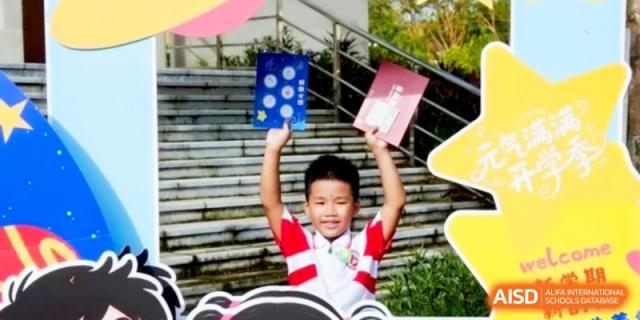 A young boy smiles brightly while holding up colorful certificates during an outdoor school celebration at RDFZ Sanya School