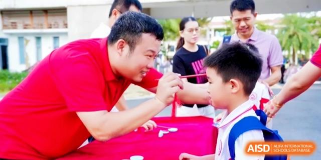 An adult in a red shirt carefully applies face paint to a young student during a festive outdoor community event at RDFZ Sanya School