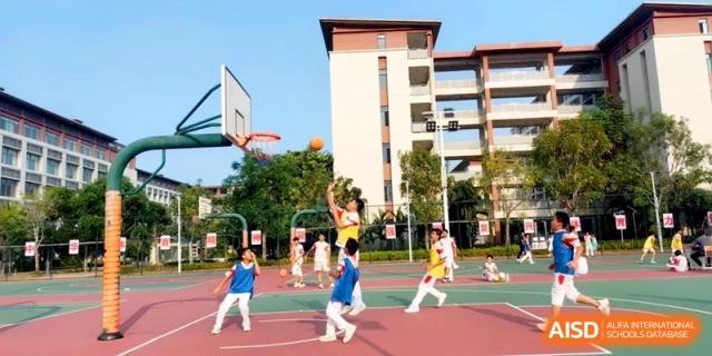 A student jumps for a layup during an active basketball game on an outdoor court surrounded by campus buildings at RDFZ Sanya School