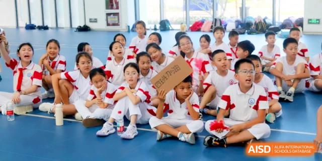 A large group of students in red and white uniforms sit on a blue indoor gym floor while listening attentively during a school assembly or activity at RDFZ Sanya School