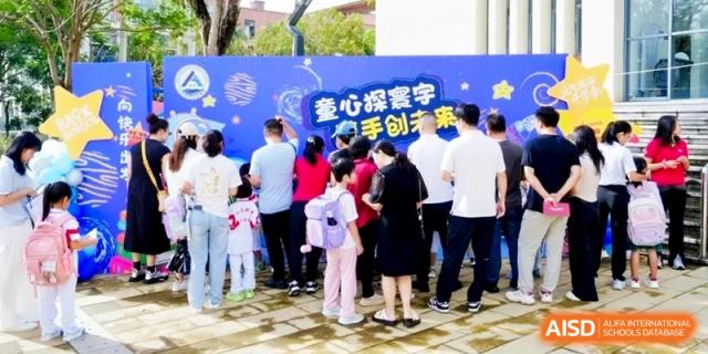 Families gather around a large blue decorative board featuring space-themed graphics and text during a vibrant outdoor community festival held at RDFZ Sanya School