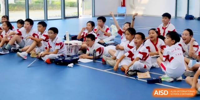 Enthusiastic students in school uniforms sit on the floor of a sports hall and raise their hands to participate in an interactive lesson or game at RDFZ Sanya School
