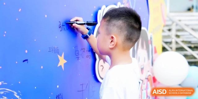 A young student focused on writing his name or a message with a black marker on a large blue themed display board during a school celebration at RDFZ Sanya School