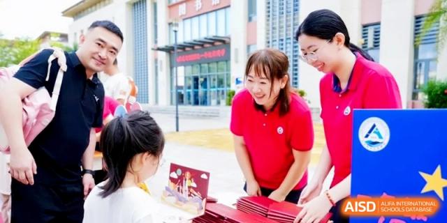 A smiling teacher in a red polo shirt interacts with a student at an outdoor booth featuring colorful books and educational materials at RDFZ Sanya School