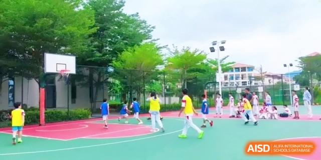 Students in athletic uniforms participate in an outdoor physical education class on a green and red sports court surrounded by lush trees at RDFZ Sanya School