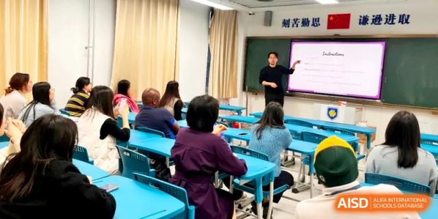 A male presenter leads a training session for a group of adults in a bright classroom at the Attached Senior School Of Shandong Normal University.