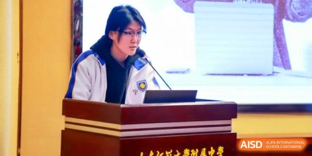A student in a school uniform presents her research using a microphone and a large screen at the Attached Senior School Of Shandong Normal University.