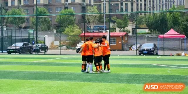 A group of football players in orange jerseys huddle together for a team talk on the field at the Attached Senior School Of Shandong Normal University.