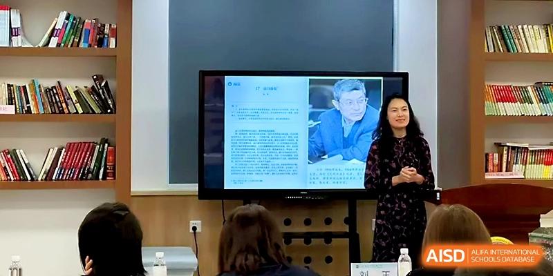 A female presenter speaks next to a monitor displaying a biographical profile in a classroom at Tianjin Foreign Languages School.