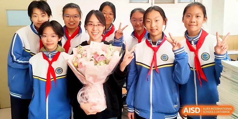 A group of students smiles for a photo while one holds a large bouquet of pink flowers at Tianjin Foreign Languages School.