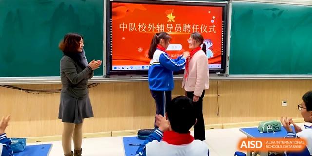 A student in uniform ties a red scarf for a female guest in front of a bright red digital backdrop at Tianjin Foreign Languages School.