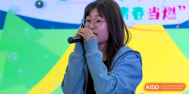 A student wearing glasses and a blue hoodie performs a vocal solo during a school talent showcase at Tianjin Foreign Languages School.
