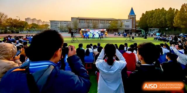 A large crowd of students gathers on the green field to watch a live performance on the outdoor stage at Tianjin Foreign Languages School.