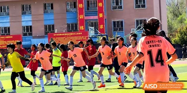 A girls' soccer team in orange uniforms competes in a match on the school's grassy field at Urumqi Bayi High School.