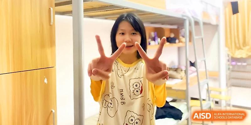 A young female student smiles and gives a peace sign while standing in her comfortable dormitory at Wuhan Guanggu Future School.