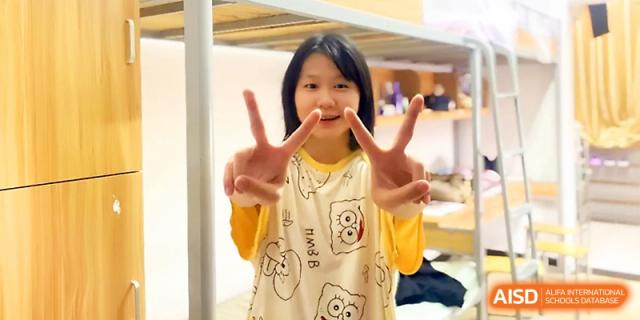 A young female student smiles and gives a peace sign while standing in her comfortable dormitory at Wuhan Guanggu Future School.