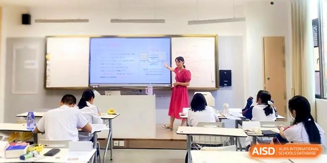 A teacher in a pink dress stands at a digital whiteboard, pointing to a lesson for her seated students at Wuhan Guanggu Future School.