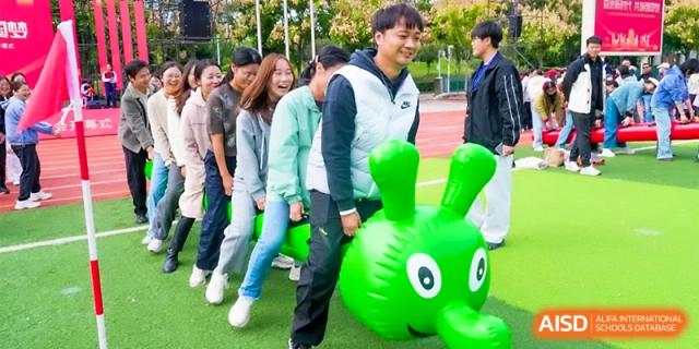 A team of staff members competes in a race using a green inflatable caterpillar toy on the grass at Wuhan Guanggu Future School.