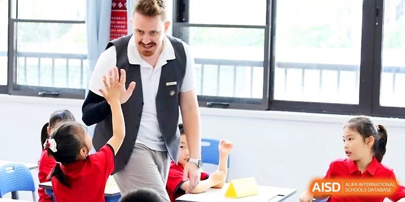 A male teacher gives a high-five to a young student in a bright, modern classroom during a primary lesson at Yung Wing School Zhuhai.