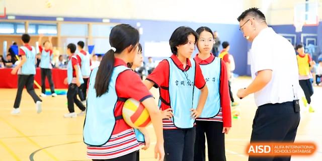 Three female students in athletic bibs hold a volleyball while waiting for their turn on the court at Yung Wing School Zhuhai.