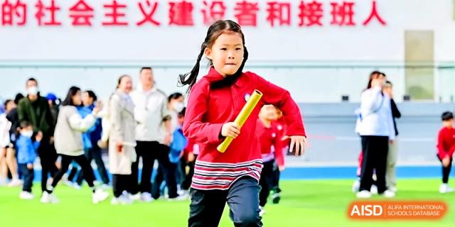 A young girl in a red athletic uniform sprints while holding a yellow relay baton during a race at Yung Wing School Zhuhai.