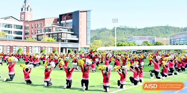 A large group of students in red outfits performs a synchronized cheer and dance routine on the grassy field at Yung Wing School Zhuhai.