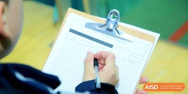 A close-up view of a hand holding a pen over a sports score sheet on a silver clipboard at Zhangjiagang Foreign Language School.