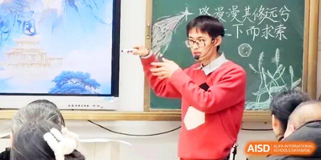 A teacher in a red sweater gestures toward a digital screen while lecturing at the front of a modern classroom at Zhengzhou No.19 High School.